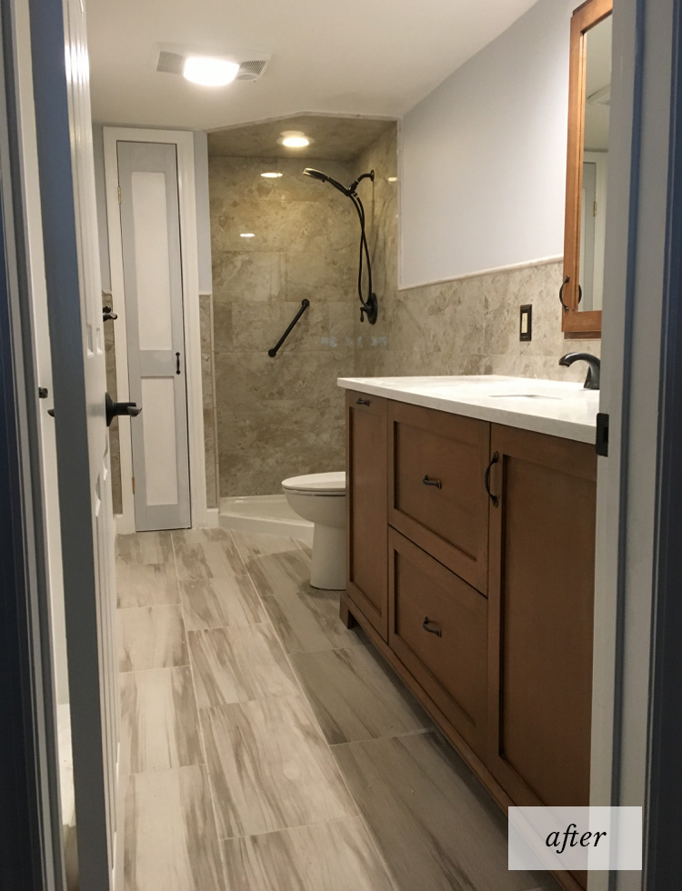 A renovated bathroom with tile floor, stand-up shower with black fixtures and a brown wood vanity sink with cabinets.