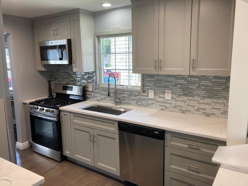 A modern style kitchen with stainless steel appliances, white cabinets and countertops and a gray and white glass tile backsplash.