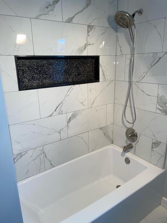 A clean white and gray marble tile shower wall with a black inset shelf and a chrome shower head.