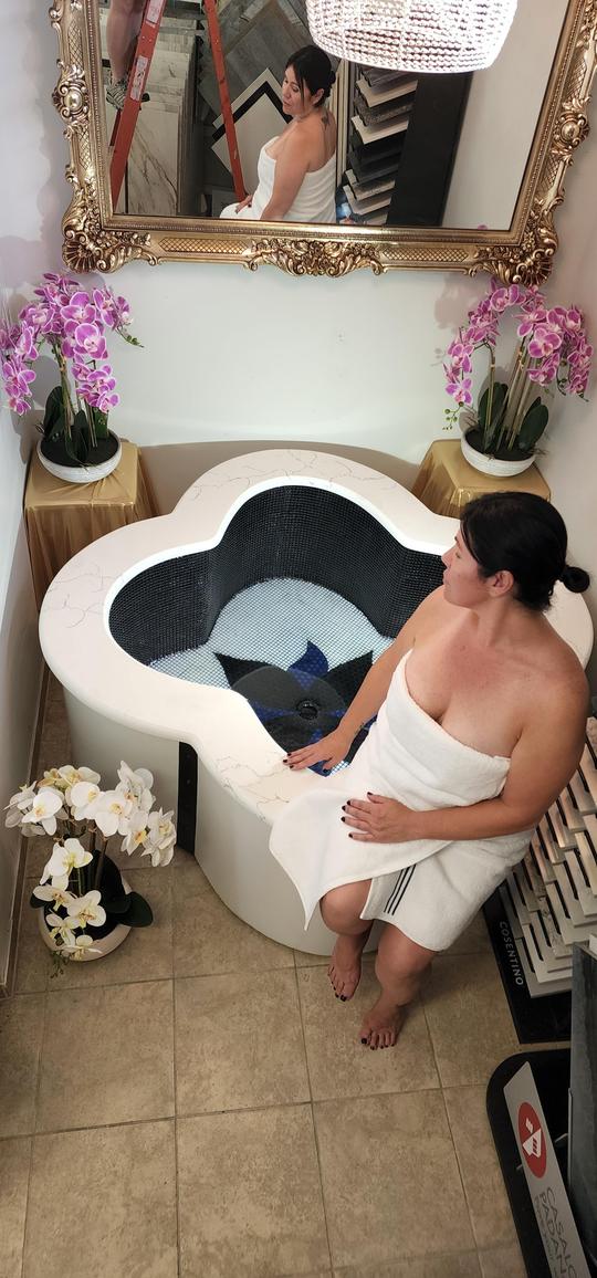 A brunette woman sitting on the ledge of a marble irregular shape bathtub staring at a pink floral arrangement on a table.